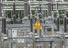 Contém um placa com aviso e um raio afixada em uma grade de arame. Ao fundo está a subestação de energia elétrica.