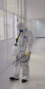Homem paramentado com máscara, luvas e avental utiliza pulverizador com solução sanitizante nos corredores de prédio